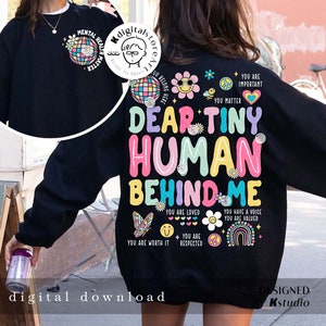 Puede incluir: Sudadera negra con un gráfico colorido que dice "Querido pequeño humano detrás de mí" con varias afirmaciones positivas e ilustraciones. El gráfico incluye un arcoíris, un signo de paz, una mariposa y flores.