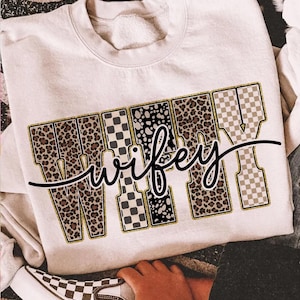 May include: Cream-coloured sweatshirt featuring the word "wifey" in a bold, decorative font. The letters are filled with leopard print, black and white polka dots, and checkered patterns. A black cursive "wifey" is written over the letters.