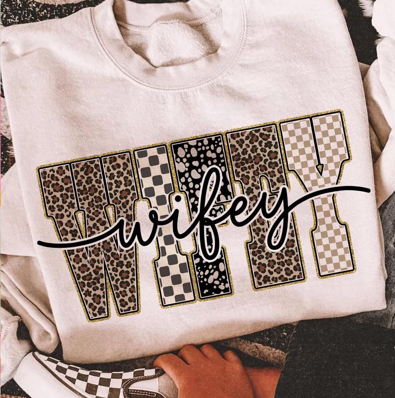 May include: Cream-coloured sweatshirt featuring the word "wifey" in a bold, decorative font. The letters are filled with leopard print, black and white polka dots, and checkered patterns. A black cursive "wifey" is written over the letters.
