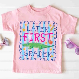 Op de afbeelding: Roze T-shirt met korte mouwen en een kleurrijke afbeelding. De afbeelding bevat de tekst "LATER FIRST GRADER" met een groene alligator, een potlood en een appel. Het ontwerp is omlijst door een blauwe rand met sterren.