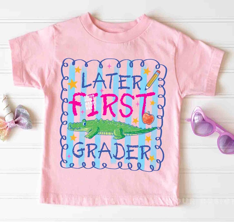 Op de afbeelding: Roze T-shirt met korte mouwen en een kleurrijke afbeelding. De afbeelding bevat de tekst "LATER FIRST GRADER" met een groene alligator, een potlood en een appel. Het ontwerp is omlijst door een blauwe rand met sterren.