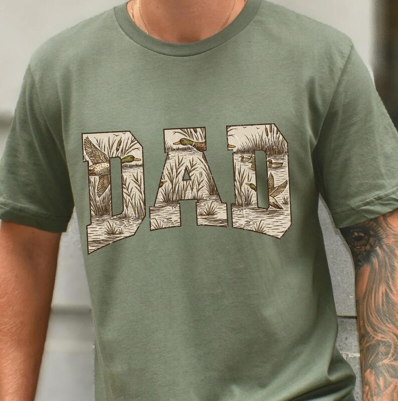May include: Olive green t-shirt with the word "DAD" in a block letter design. The letters are filled with a detailed illustration of ducks, water, and reeds, creating a nature-themed graphic.
