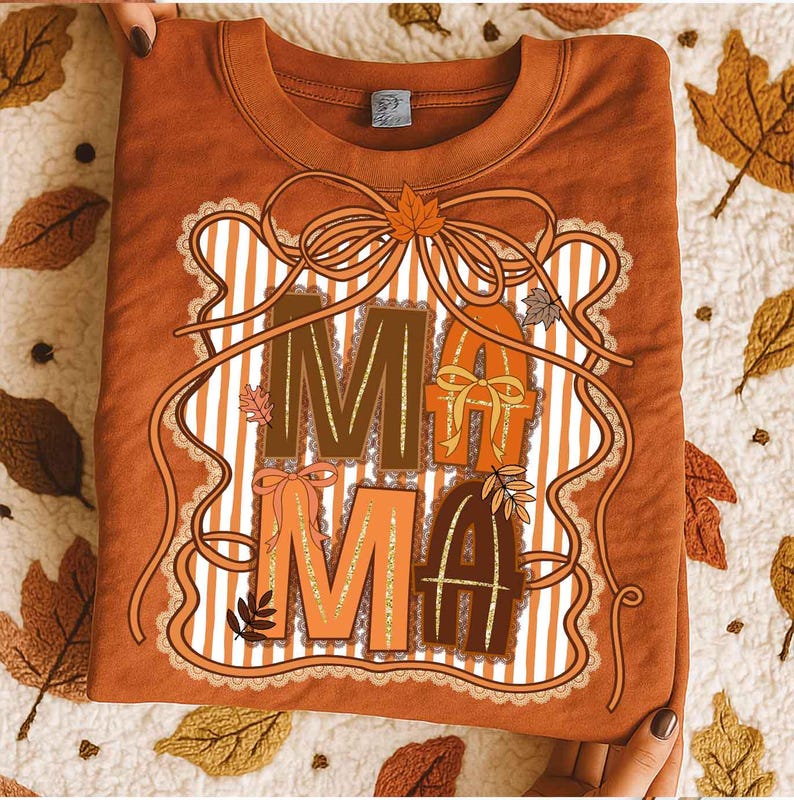 Op de afbeelding: Een roestkleurig T-shirt met het woord "MAMA" in een herfstthema-ontwerp. De letters zijn in bruin- en oranje tinten, met decoratieve strikken en bladeren. Het ontwerp wordt omlijst door een wit-oranje gestreepte achtergrond en een kantachtige rand.