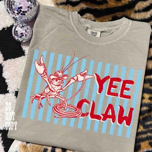 May include: A gray t-shirt with a red and blue graphic. The graphic features a cartoon crawfish wearing a cowboy hat and holding a lasso, with the words "Yee Claw" in red letters.