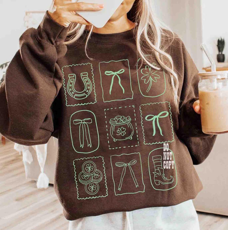 May include: A brown sweatshirt with green illustrations of a horseshoe, bows, a four-leaf clover, a pot of gold, and coins. The text "DO NOT COPY" is also visible. The sweatshirt has long sleeves.