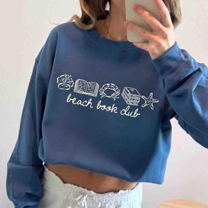 May include: A blue sweatshirt with the words "beach book club" in white script. The design includes white line drawings of a flower, an open book, a crab, a stack of books, and a starfish. The sweatshirt has long sleeves.