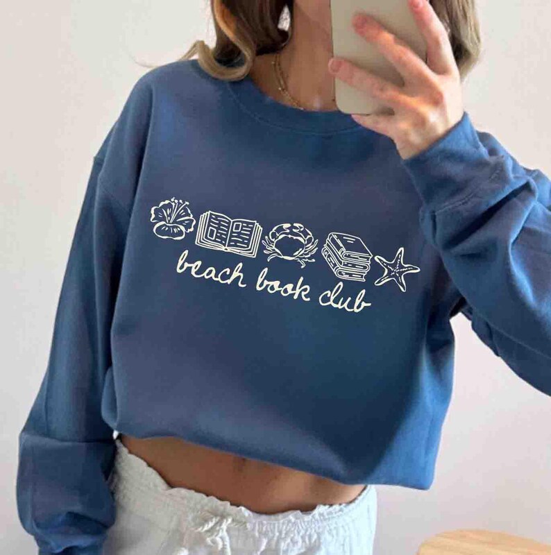 May include: A blue sweatshirt with the words "beach book club" in white script. The design includes white line drawings of a flower, an open book, a crab, a stack of books, and a starfish. The sweatshirt has long sleeves.