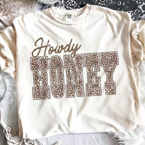 May include: Cream-colored t-shirt with the words "Howdy HONEY" in a brown, glittery font. The word "HONEY" is filled with a leopard print pattern. The shirt is a casual style, perfect for everyday wear.