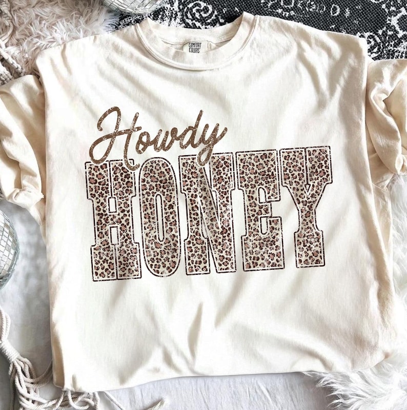 May include: Cream-colored t-shirt with the words "Howdy HONEY" in a brown, glittery font. The word "HONEY" is filled with a leopard print pattern. The shirt is a casual style, perfect for everyday wear.