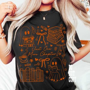 May include: Black t-shirt with orange graphic illustrations of books, ghosts, bows, and stars. The text "Just One More Chapter" is also in orange. The shirt is a comfortable, casual style.