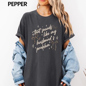 May include: Dark gray t-shirt with the text "that sounds like my husband's problem" in a decorative font. The text is accented with star and floral designs. The shirt is part of a casual wear collection.