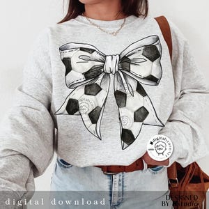 May include: A gray sweatshirt with a black and white soccer ball bow design. The bow is tied in a knot and the ends of the bow are made of soccer balls. The sweatshirt has a white background and the bow is in the center of the sweatshirt.
