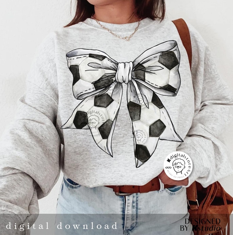 May include: A gray sweatshirt with a black and white soccer ball bow design. The bow is tied in a knot and the ends of the bow are made of soccer balls. The sweatshirt has a white background and the bow is in the center of the sweatshirt.