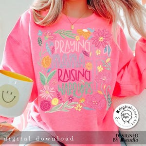 May include: Pink sweatshirt with a floral design and the text "Praying Mama Raising Warriors".