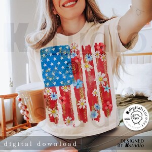 Puede incluir: Una camiseta blanca con un dise&ntilde;o de bandera estadounidense roja, blanca y azul. La bandera est&aacute; pintada con un efecto acuarela y tiene margaritas blancas esparcidas por toda ella. El texto "Kstudio" es visible en el dise&ntilde;o.