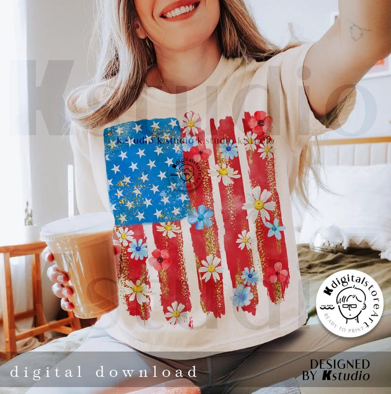 Puede incluir: Una camiseta blanca con un dise&ntilde;o de bandera estadounidense roja, blanca y azul. La bandera est&aacute; pintada con un efecto acuarela y tiene margaritas blancas esparcidas por toda ella. El texto "Kstudio" es visible en el dise&ntilde;o.