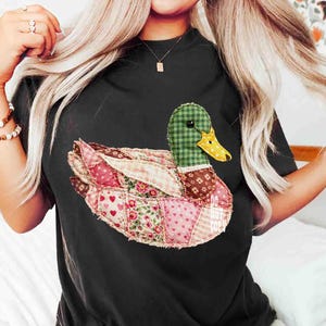 May include: Black t-shirt featuring a patchwork duck design. The duck has a green checkered head, a yellow beak, and wings made of various patterned fabrics, including pink hearts and floral prints. The text "DO NOT COPY" is visible.