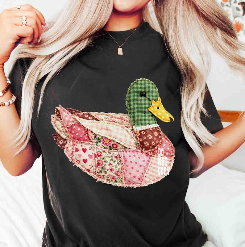 May include: Black t-shirt featuring a patchwork duck design. The duck has a green checkered head, a yellow beak, and wings made of various patterned fabrics, including pink hearts and floral prints. The text "DO NOT COPY" is visible.