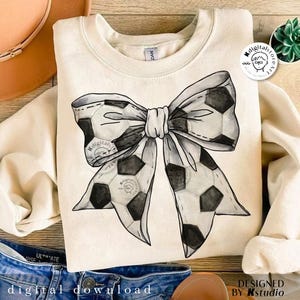 May include: A white sweatshirt with a black and white watercolor design of a soccer ball bow. The design is centered on the sweatshirt.