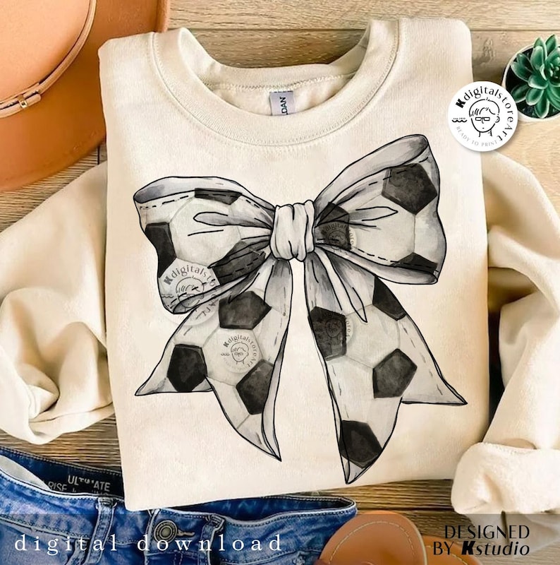 May include: A white sweatshirt with a black and white watercolor design of a soccer ball bow. The design is centered on the sweatshirt.