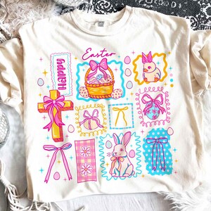 May include: Cream-colored t-shirt with a colorful Easter-themed graphic. The design includes a cross, a basket of eggs, a bunny, and the word "Easter" in pink script. Other elements include bows, eggs, and the word "Happy".