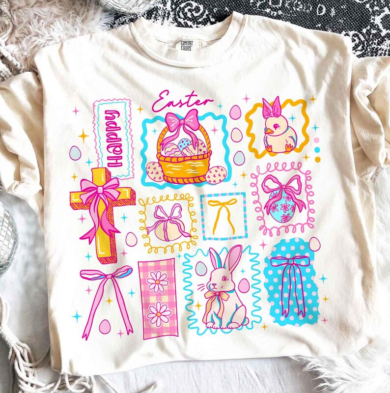 May include: Cream-colored t-shirt with a colorful Easter-themed graphic. The design includes a cross, a basket of eggs, a bunny, and the word "Easter" in pink script. Other elements include bows, eggs, and the word "Happy".