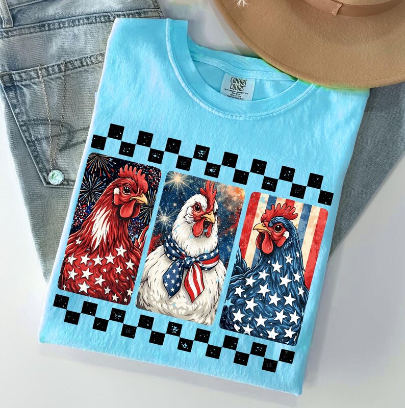 May include: A light blue t-shirt featuring three chickens with patriotic designs. The chickens are in red, white, and blue, with stars and stripes. The shirt is folded on top of a pair of blue jeans and a straw hat.