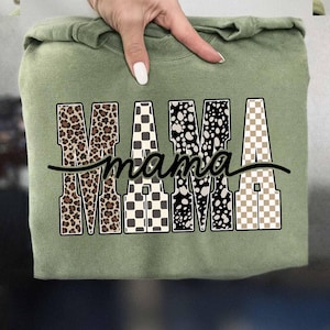 May include: Sage green sweatshirt with the word "MAMA" in a bold, patterned design. The letters feature leopard print, checkered, and polka dot patterns. A black cursive "mama" is written underneath the letters.
