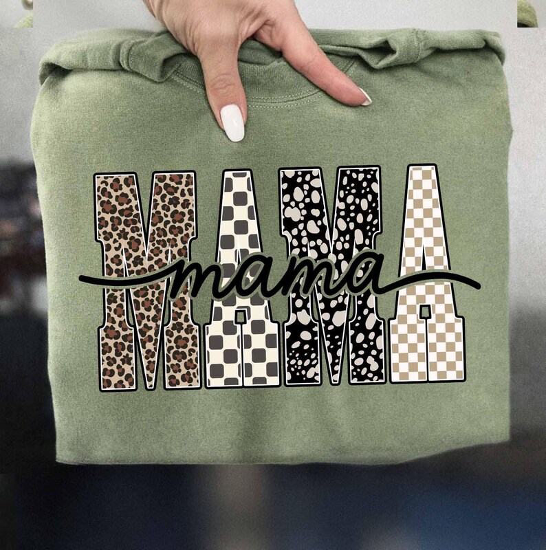 May include: Sage green sweatshirt with the word "MAMA" in a bold, patterned design. The letters feature leopard print, checkered, and polka dot patterns. A black cursive "mama" is written underneath the letters.