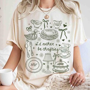 May include: Cream-colored t-shirt with a green illustration of teacups, plates, and a duck, along with the text "I'd rather be thrifting." The design includes floral and bow accents, creating a vintage aesthetic.