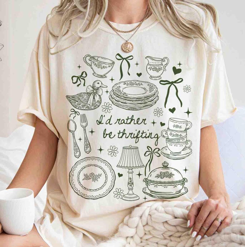 May include: Cream-colored t-shirt with a green illustration of teacups, plates, and a duck, along with the text "I'd rather be thrifting." The design includes floral and bow accents, creating a vintage aesthetic.