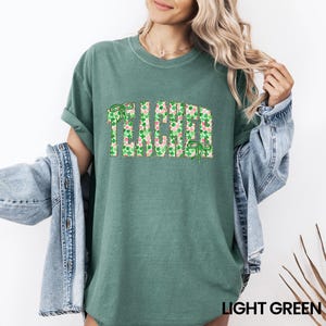 May include: Light green t-shirt with the word "TEACHER" in a green and pink shamrock pattern. The shirt has rolled-up sleeves and a crew neck. The text "LIGHT GREEN" is at the bottom of the image.