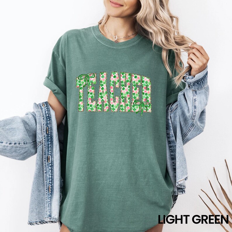 May include: Light green t-shirt with the word "TEACHER" in a green and pink shamrock pattern. The shirt has rolled-up sleeves and a crew neck. The text "LIGHT GREEN" is at the bottom of the image.