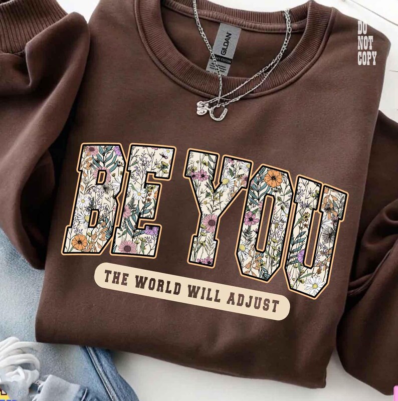 May include: A brown sweatshirt with the words "BE YOU" in a floral design. Below the text is the phrase "THE WORLD WILL ADJUST" in a white banner. A silver necklace is draped on the sweatshirt.