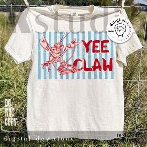 May include: Cream-colored t-shirt with a red cartoon crawfish wearing a cowboy hat and holding a lasso. The text "YEE CLAW" is in red, with blue vertical stripes in the background. The shirt is hanging on a clothesline.