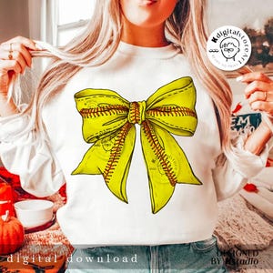 Puede incluir: Una sudadera blanca con un dise&ntilde;o de lazo de softball amarillo y rojo. El lazo est&aacute; atado en un nudo y los extremos del lazo fluyen. La sudadera es un gran regalo para los jugadores de softball.