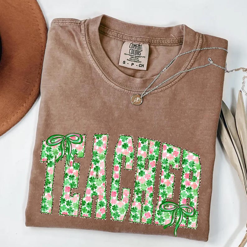 May include: A brown Comfort Colors t-shirt with the word "TEACHER" in green and pink shamrock pattern letters. The shirt has a crew neck and short sleeves. A silver necklace with a small pendant is on the shirt.