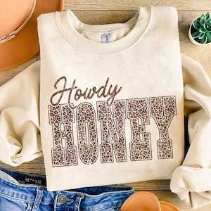 May include: Cream-colored sweatshirt with the text "Howdy HONEY" in brown leopard print. The sweatshirt is folded, with a pair of blue jeans, a brown hat, and sandals in the background. A small succulent plant is also visible.