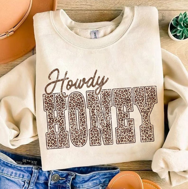 May include: Cream-colored sweatshirt with the text "Howdy HONEY" in brown leopard print. The sweatshirt is folded, with a pair of blue jeans, a brown hat, and sandals in the background. A small succulent plant is also visible.