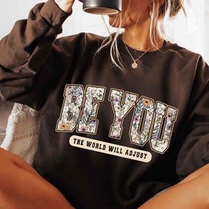 May include: Dark brown sweatshirt with the words "BE YOU" in large, floral-filled letters. Below, the text reads "THE WORLD WILL ADJUST" in a smaller, rectangular banner. The sweatshirt is a casual style.