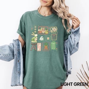 May include: Light green t-shirt featuring a colorful Easter-themed graphic. The design includes framed illustrations of a bunny, eggs, carrots, and other spring elements. The text "LIGHT GREEN" is printed at the bottom.