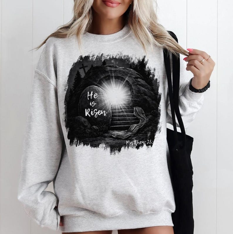 May include: Light gray sweatshirt featuring a black and white graphic of an open tomb with a bright light emanating from within. The text "He is Risen" is displayed on a stone, with "Matthew 28" below.