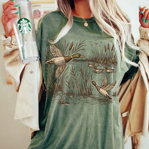 May include: Green t-shirt featuring a detailed illustration of ducks in a marsh setting. The design includes ducks in flight and swimming, with reeds and water. The shirt is a muted green color.