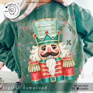 May include: A green crewneck sweatshirt featuring a central graphic of a nutcracker wearing a teal and gold crown, red jacket with gold trim, and white hair. The design is surrounded by green pine branches, red berries, and pink snowflakes.