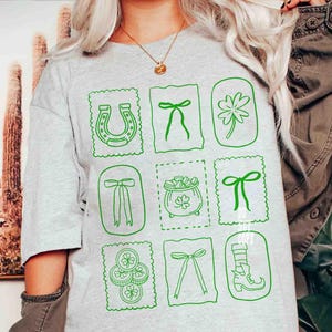 May include: Light gray t-shirt featuring green St. Patrick's Day themed graphics. The design includes a horseshoe, bows, a four-leaf clover, a pot of gold, and a leprechaun boot. The graphics are framed in a postage stamp style.