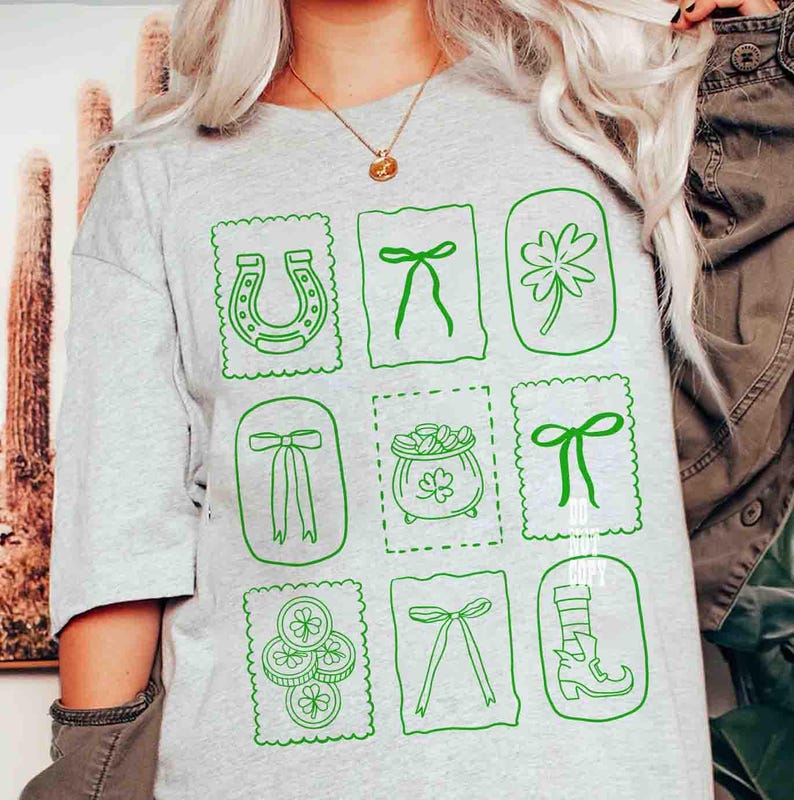 May include: Light gray t-shirt featuring green St. Patrick's Day themed graphics. The design includes a horseshoe, bows, a four-leaf clover, a pot of gold, and a leprechaun boot. The graphics are framed in a postage stamp style.