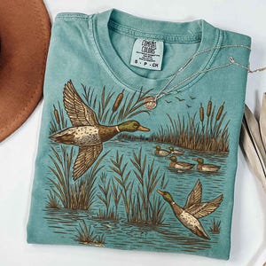 May include: A teal Comfort Colors t-shirt featuring a detailed illustration of ducks in a marsh setting. The design includes ducks in flight and swimming, cattails, and water. A silver necklace with a pendant rests on the shirt.