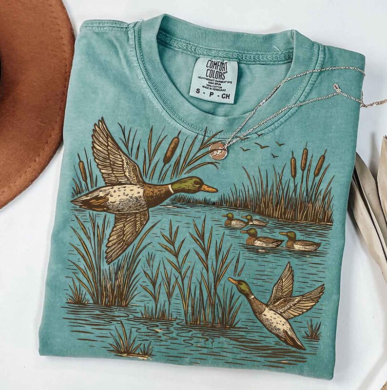 May include: A teal Comfort Colors t-shirt featuring a detailed illustration of ducks in a marsh setting. The design includes ducks in flight and swimming, cattails, and water. A silver necklace with a pendant rests on the shirt.