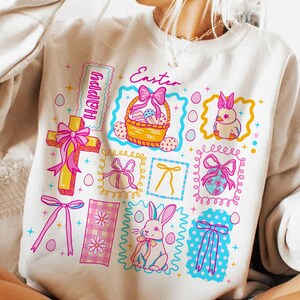 May include: Cream-colored sweatshirt with a colorful Easter-themed graphic. The design includes a cross, a basket of eggs, a bunny, and the word "Happy" and "Easter". The graphic is framed with pink, yellow, and blue accents.