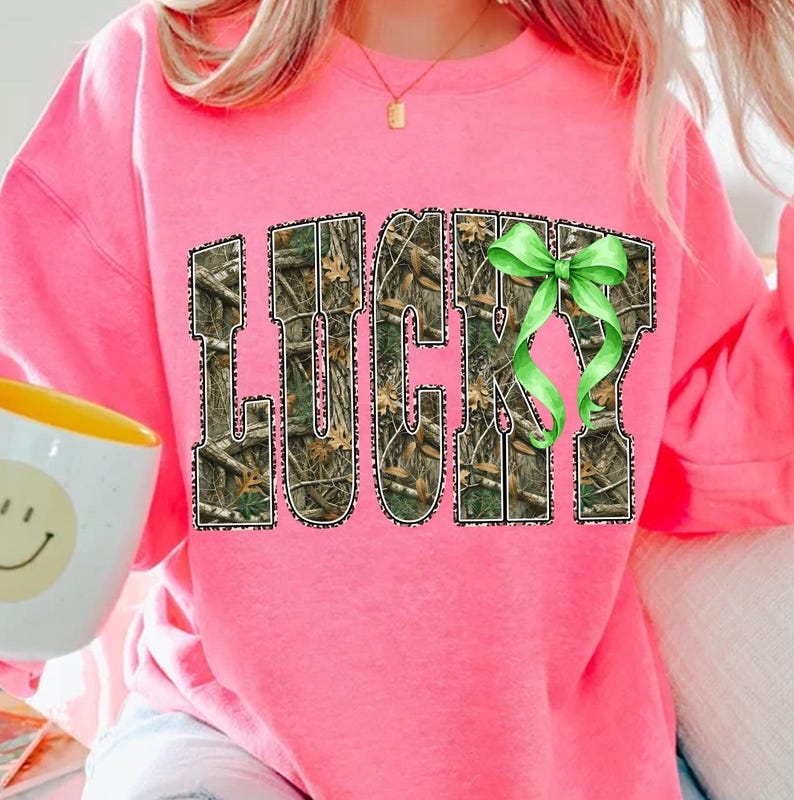 May include: A bright pink sweatshirt with the word "LUCKY" in a camouflage pattern with a green bow. The sweatshirt is a crew neck style and is made of a soft material. The image also shows a white mug with a yellow interior.